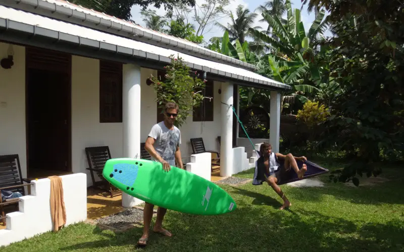 A guest carrying a green soft-top surfboard across the garden in front of the rooms at Surfing Life